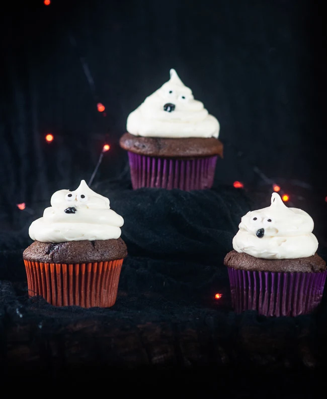 Three ghost cupcakes on a black background with orange twinkle lights make a bootiful treat for Halloween.