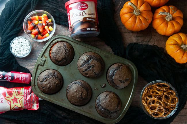 Ingredients for spider cupcakes: chocolate cupcakes, candy corn, candy eyes, chocolate frosting and pretzels on black with pumpkins