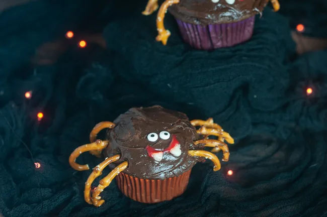 A chocolate cupcake decorated like a spider with pretzel legs and candy eyes for Halloween on black