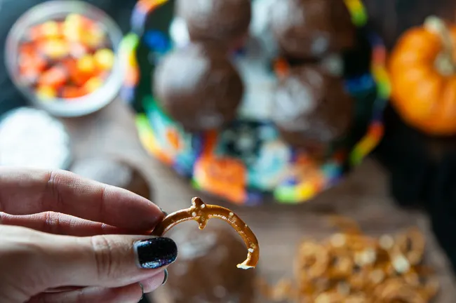 A hand holds a curved piece of a pretzel for the spider's leg above a plate of chocolate cupcakes