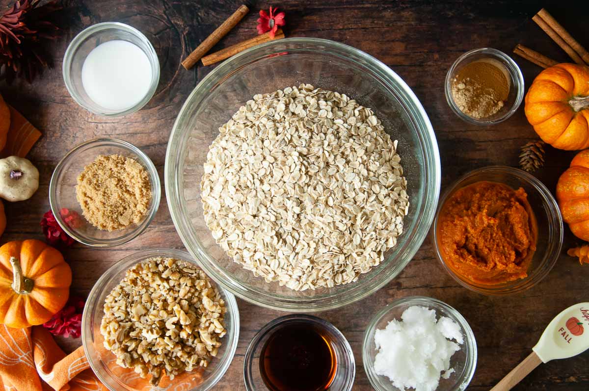 Glass bowls with the ingredients for pumpkin granola (milk, walnuts, pumpkin, coconut oil, pumpkin puree, spices and rolled oats) sit on a wooden table