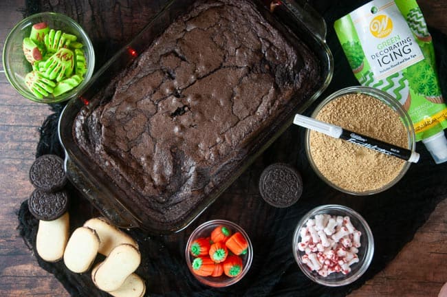 Ingredients for graveyard brownies are brownies, Milanos, cookie and graham cracker crumbs, bone shaped sprinkles, a food writer, candy pumpkins and edible zombie hands. All shown on a wood table with black fabric.