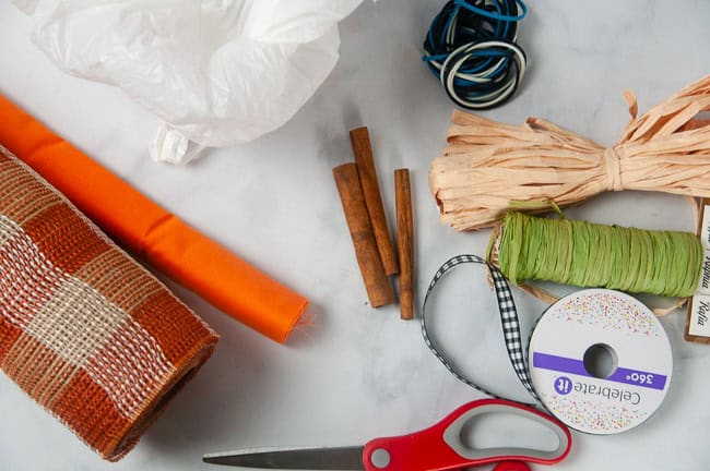 Material for Easy Fabric Pumpkins include fabric or burlap, scissors, cinnamon sticks, plastic bags, rubber bands, raffia, ribbon and other embellishments