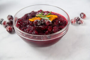 A clear bowl of Instant Pot Cranberry Sauce surrounded by cranberries on white topped with an orange slice and rosemary.