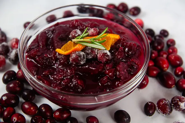 A clear bowl of Instant Pot Cranberry Sauce surrounded by cranberries on white topped with an orange slice and rosemary.