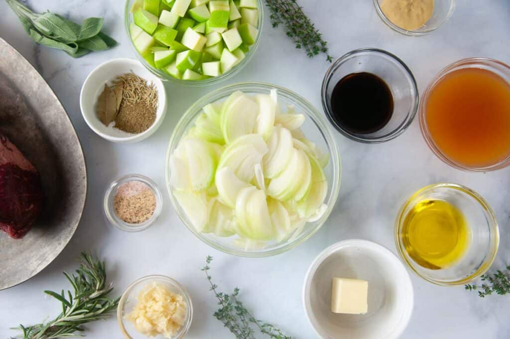 The ingredients for Instant Pot Roast Beef: chuck roast, onions, apples, balsamic vinaigrette, garlic powder, Dijon mustard, butter, olive oil, salt, pepper, apple cider, bay leaves, sage, rosemary, and fresh garlic.