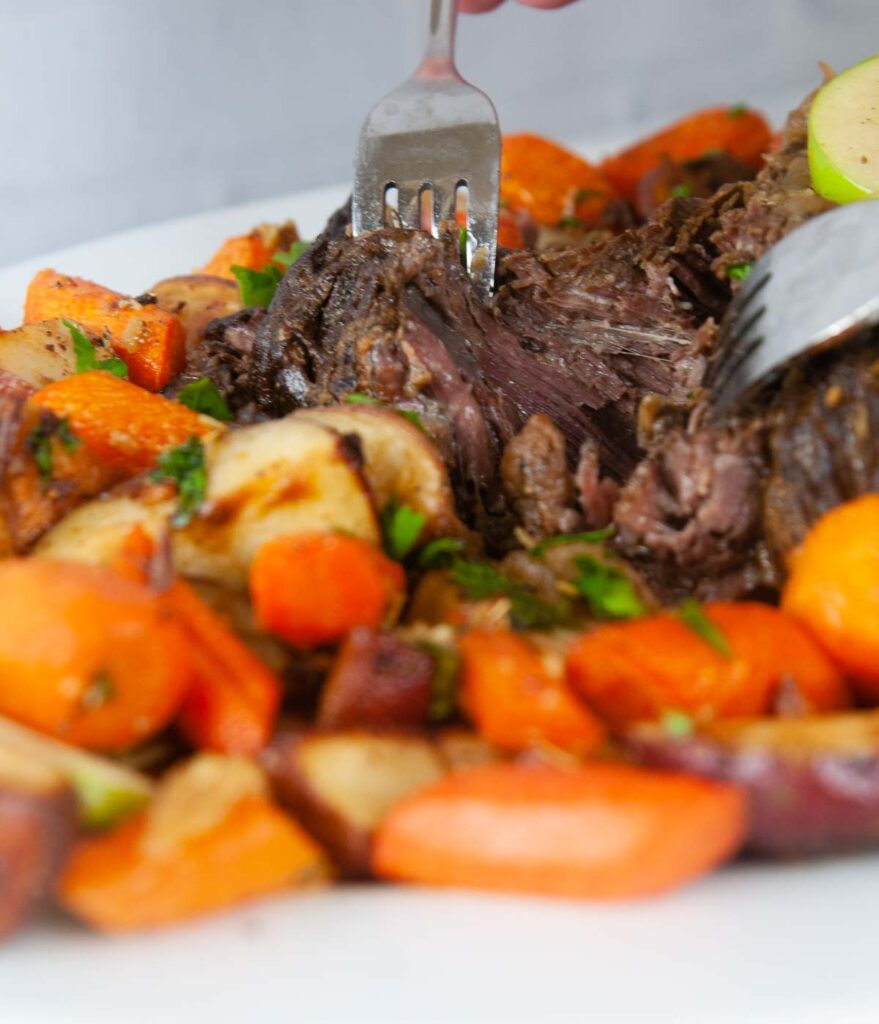 A forkful of tender Instant Pot Roast beef is slathered in apple cider gravy and served with roasted carrots and potatoes.