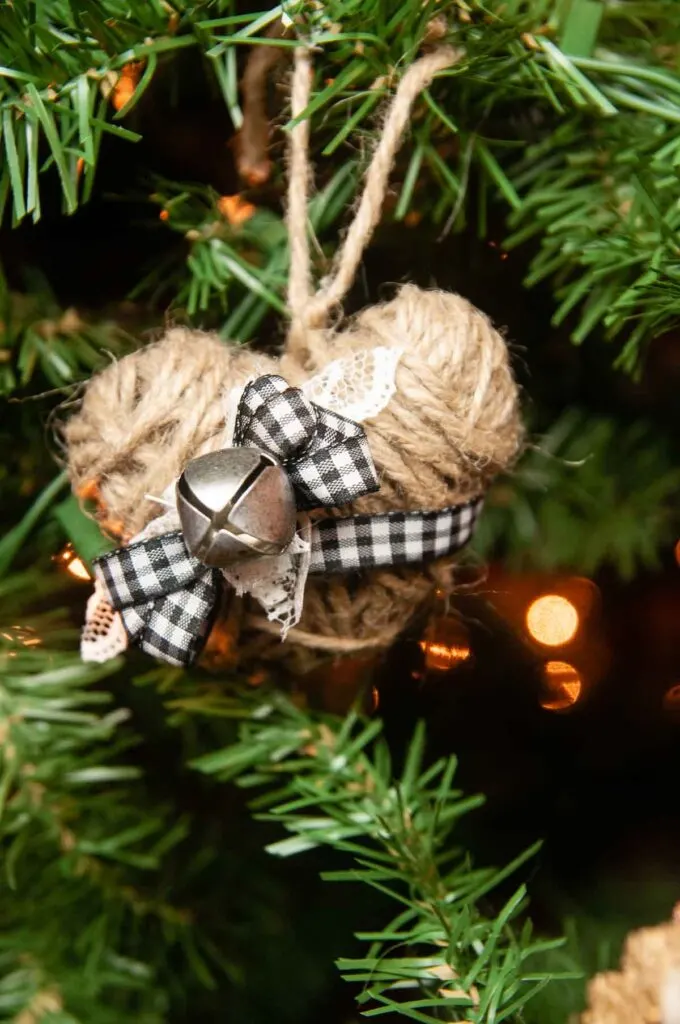 A twine heart decorated for Christmas with a jingle bell and buffalo plaid bow on a Christmas tree.