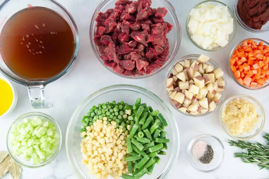 Ingredients for vegetable beef soup shown in glass bowls. Ingredients include: olive oil, beef broth, celery, carrots, onions, potatoes, beef, tomato paste and spices.