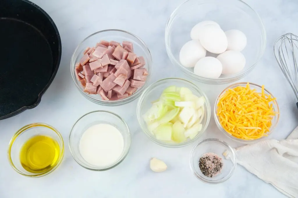 Ingredients for ham and cheese frittata on a white counter in bowls. Ingredients: chopped ham, heavy cream, olive oil, eggs, onion, cheese, garlic, salt and pepper