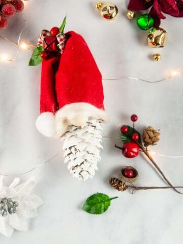 Easy pinecone gnomes are a quick adorable diy gnome ornament. A painted pinecone wears a red felt hat with a puff ball and bow so it looks like a Christmas gnome.