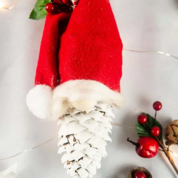 Easy pinecone gnomes are a quick adorable diy gnome ornament. A painted pinecone wears a red felt hat with a puff ball and bow so it looks like a Christmas gnome.