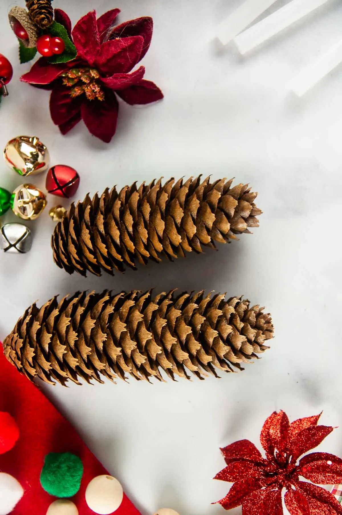 Materials to make a pinecone gnome ornament include long pinecones, white paint, felt, wood beads, hot glue sticks, and other decorations