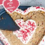 A piece cut out of a heart shaped chocolate chip cookie cake