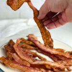 A hand picking up a crispy piece of bacon off a plate